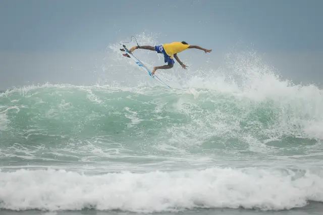 Surfer in a yellow shirt above a breaking wave, with spray behind the board.